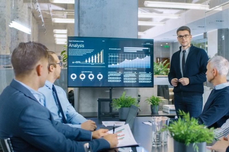 Young Stock Trader Shows to the Executive Managers Cryptocurrency and Trade Market Correlation Pointing at the Wall TV.